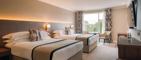 A hotel room with two beds, a desk, a chair, and a large window with curtains.