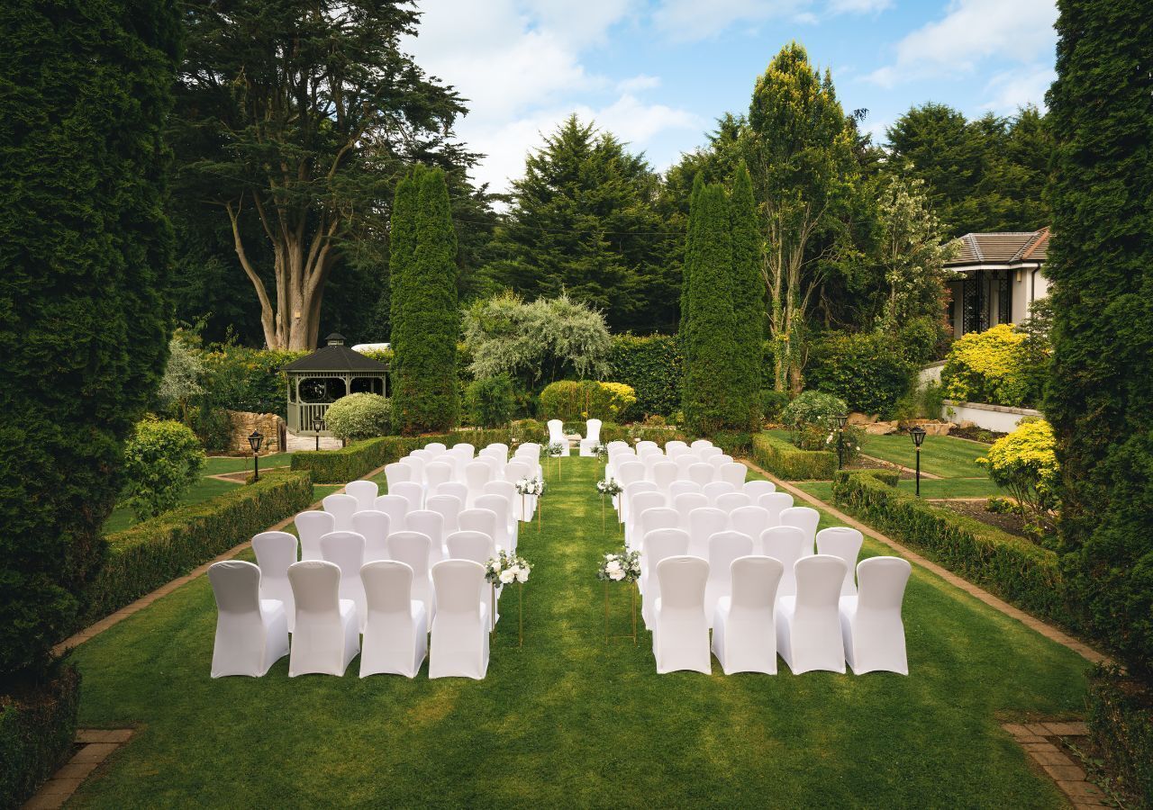 A formal outdoor setting with rows of white chairs arranged for an event, surrounded by lush greenery and a clear sky.