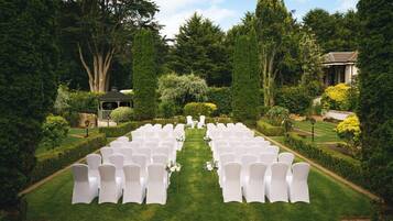 A formal outdoor setting with rows of white chairs arranged for an event, surrounded by lush greenery and a clear sky.