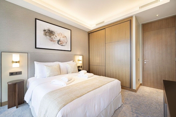 A hotel room with a large bed, bedside lamps, a framed artwork, a wardrobe, and a desk.