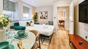 Een moderne hotelkamer met een bed, een zithoek met een tafel gedekt voor twee personen, een groene bank en een groot raam met gordijnen.