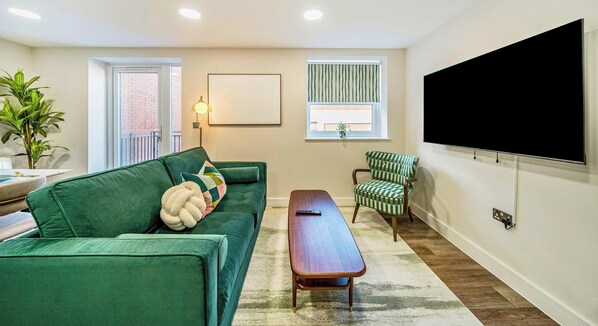 A living room with a green sofa, a wooden coffee table, a checkered armchair, a large TV, and a window with striped curtains. - Town Center| Contractors/Families | WiFi | Balcony (Aldershot)