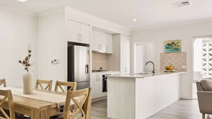 A modern kitchen with white cabinetry, a central island, and a dining area with a wooden table and chairs. - Quenda Villa (Canning Vale)