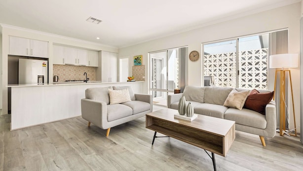 A modern living room with a sofa, coffee table, and a kitchen area with cabinets and a refrigerator. - Quenda Villa (Canning Vale)