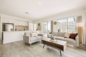 A modern living room with a sofa, coffee table, and a kitchen area with cabinets and a refrigerator. - Quenda Villa (Canning Vale)
