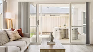 A modern living room with a sofa, a coffee table, and a sliding glass door leading to an outdoor area with a patio and a view of a building. - Quenda Villa (Canning Vale)