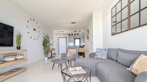 Ein modernes Wohnzimmer mit einem grauen Sofa, einem Glastisch und einem an der Wand montierten Fernseher. Üchtöpfe und ein Bücherregal mit dekorativen Accessoires sind ebenfalls vorhanden.