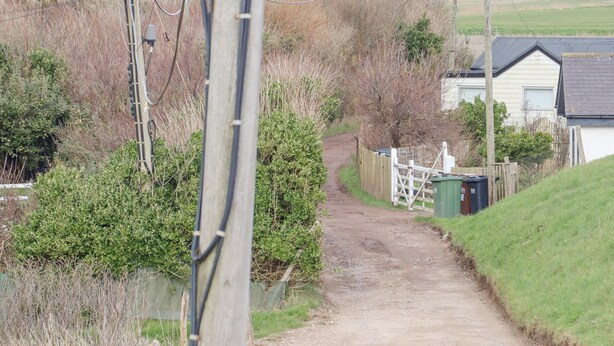 A rural dirt path leading to a house with a white fence and a green outbuilding.