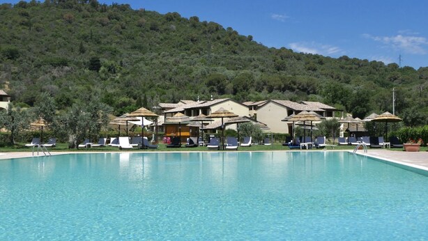 Una piscina con sillas de descanso y sombrillas, rodeada de edificios con techos de paja, ubicada al pie de una colina.