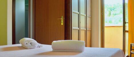 A hotel room with a bed, a folded towel, a wardrobe, and a sliding glass door leading to an outdoor area.