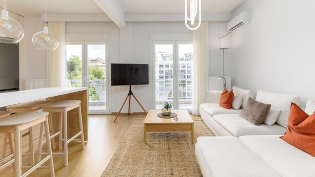 A modern living room with a white sofa, a coffee table, and a TV. There are wooden stools and a large window with a view of buildings. - Bel Air by halu (Thessaloniki)