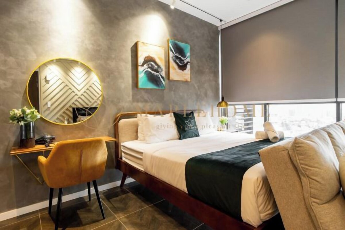 A modern hotel room with a bed, a desk, and a chair. There are two paintings on the wall and a round mirror above the desk.