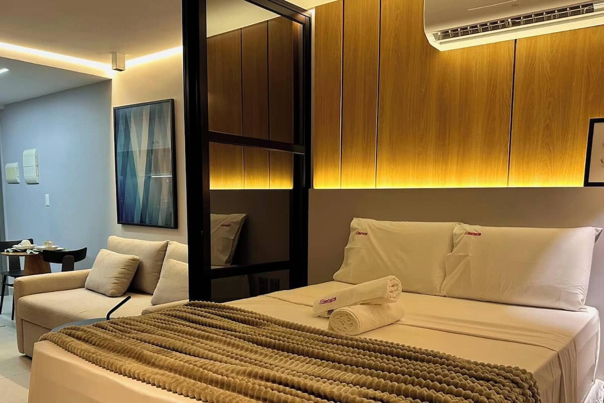 A hotel room with a bed, a desk, a chair, a painting, and an air conditioning unit.