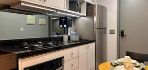 A compact kitchen with a gas stove, a coffee machine, a refrigerator, and a small dining table with plates and cups. - Vert House - LikeHome Hospedagens (João Pessoa)