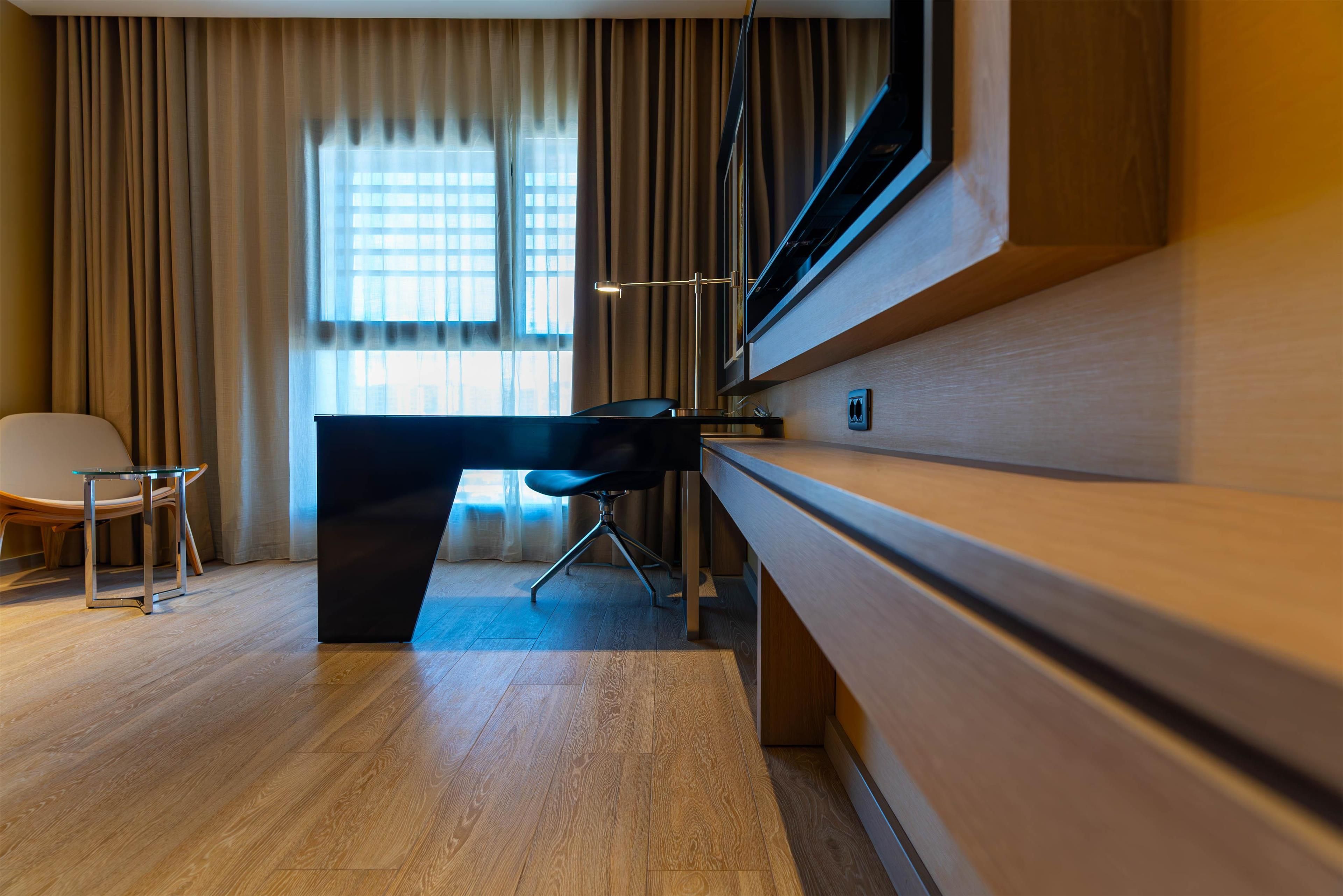 Habitación de hotel moderna con un escritorio de madera, una silla, un ventanal con cortinas y un televisor de pantalla plana montado en la pared.