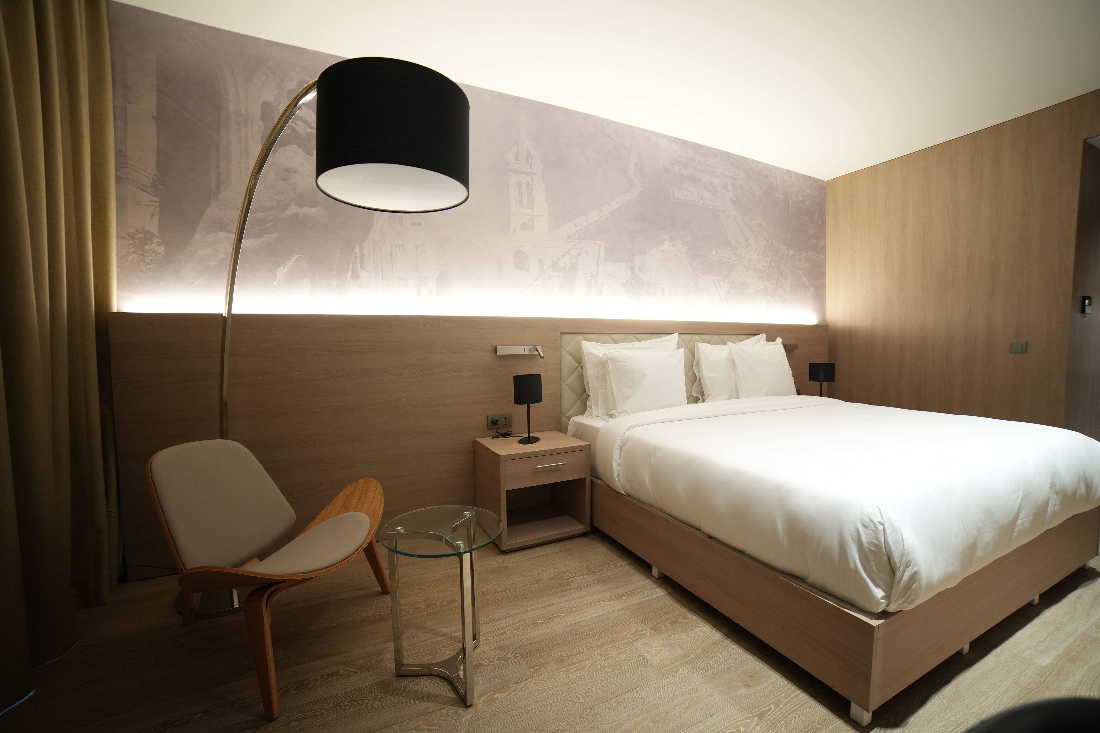 A modern hotel room with a large bed, a nightstand with a lamp, a glass table, and a chair.