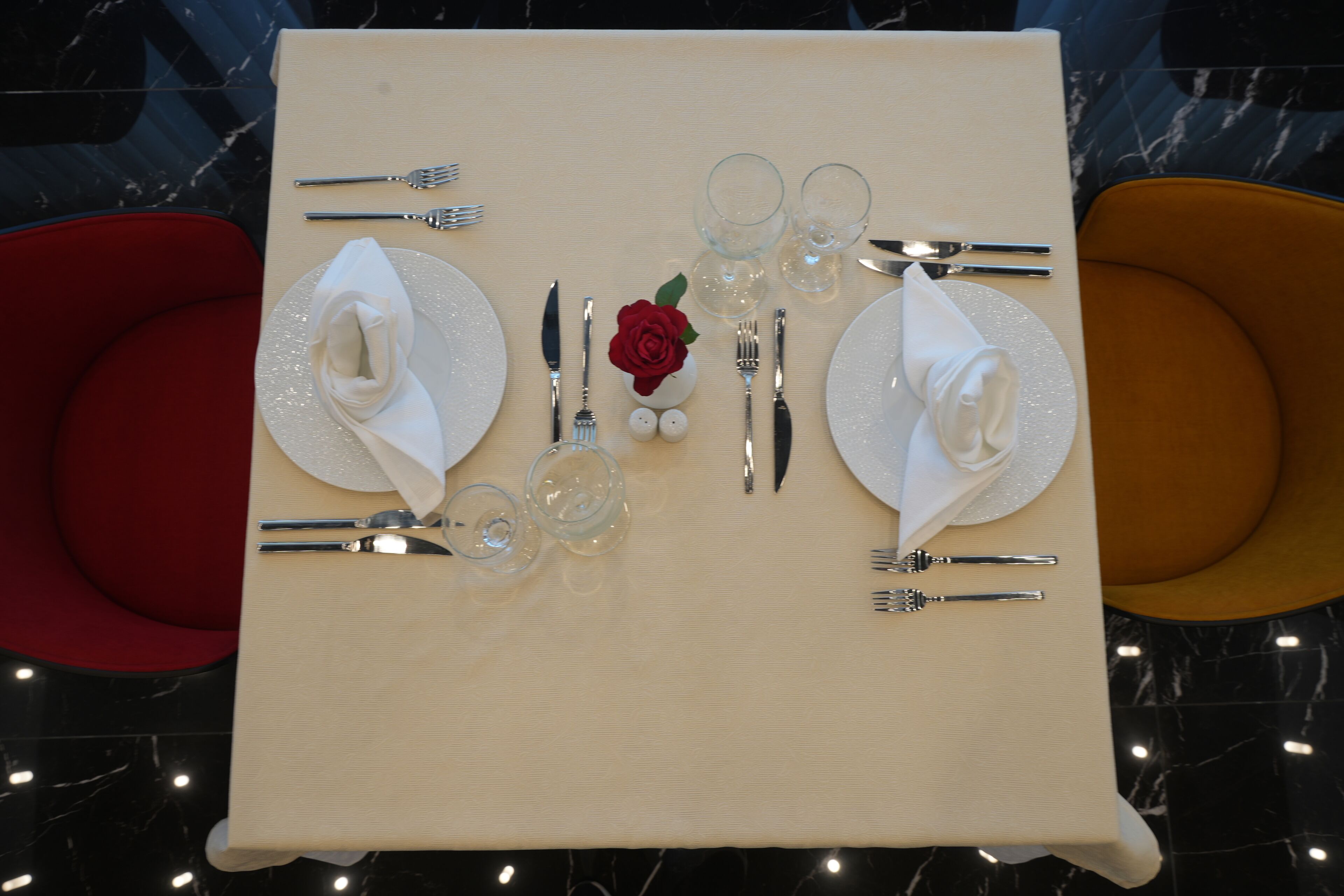 A table set for two with a white tablecloth, silverware, clear glasses, and a red rose centerpiece.