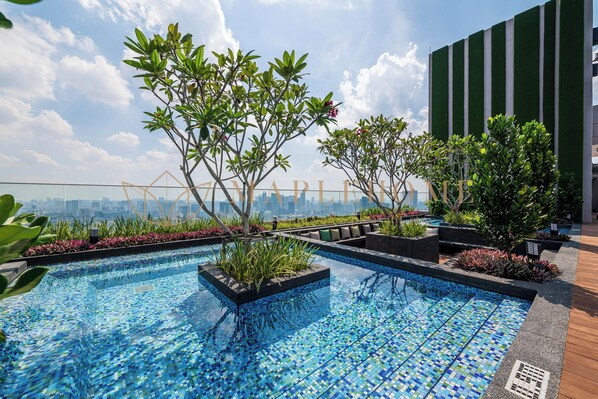 Una piscina en la azotea con mosaicos, rodeada de macetas con diversas plantas y un horizonte urbano de fondo.