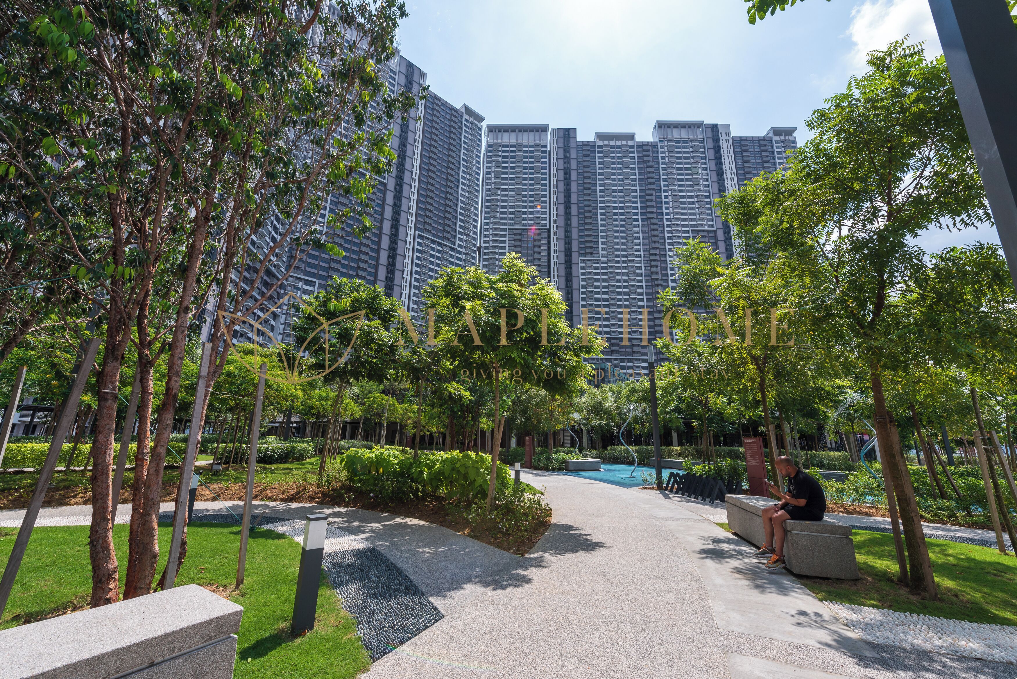 A pathway lined with trees and a bench, leading to a modern building complex with high-rise structures in the background.