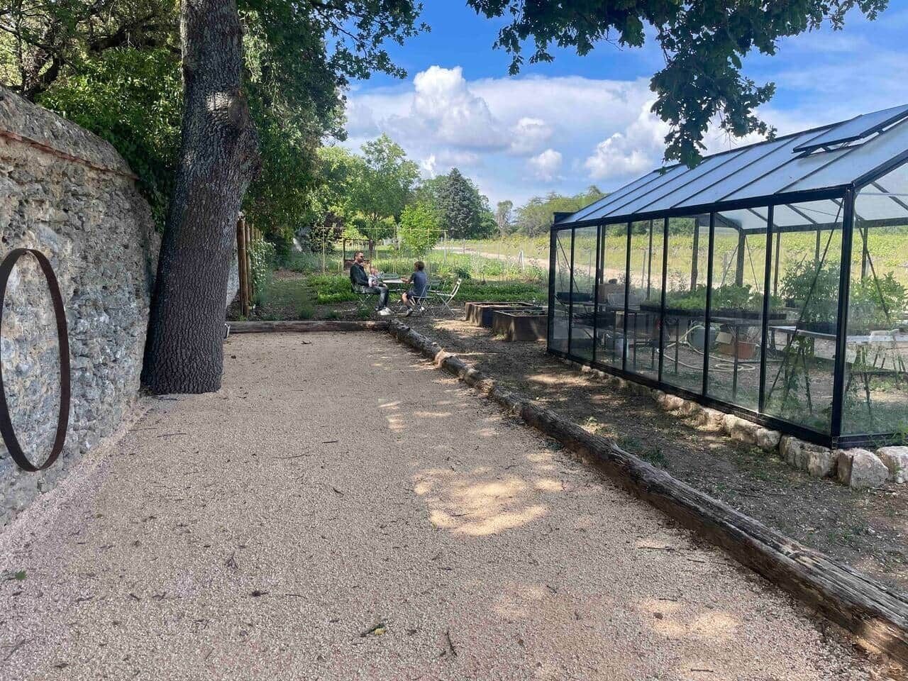 A greenhouse with a gravel path, a stone wall, and a tree.