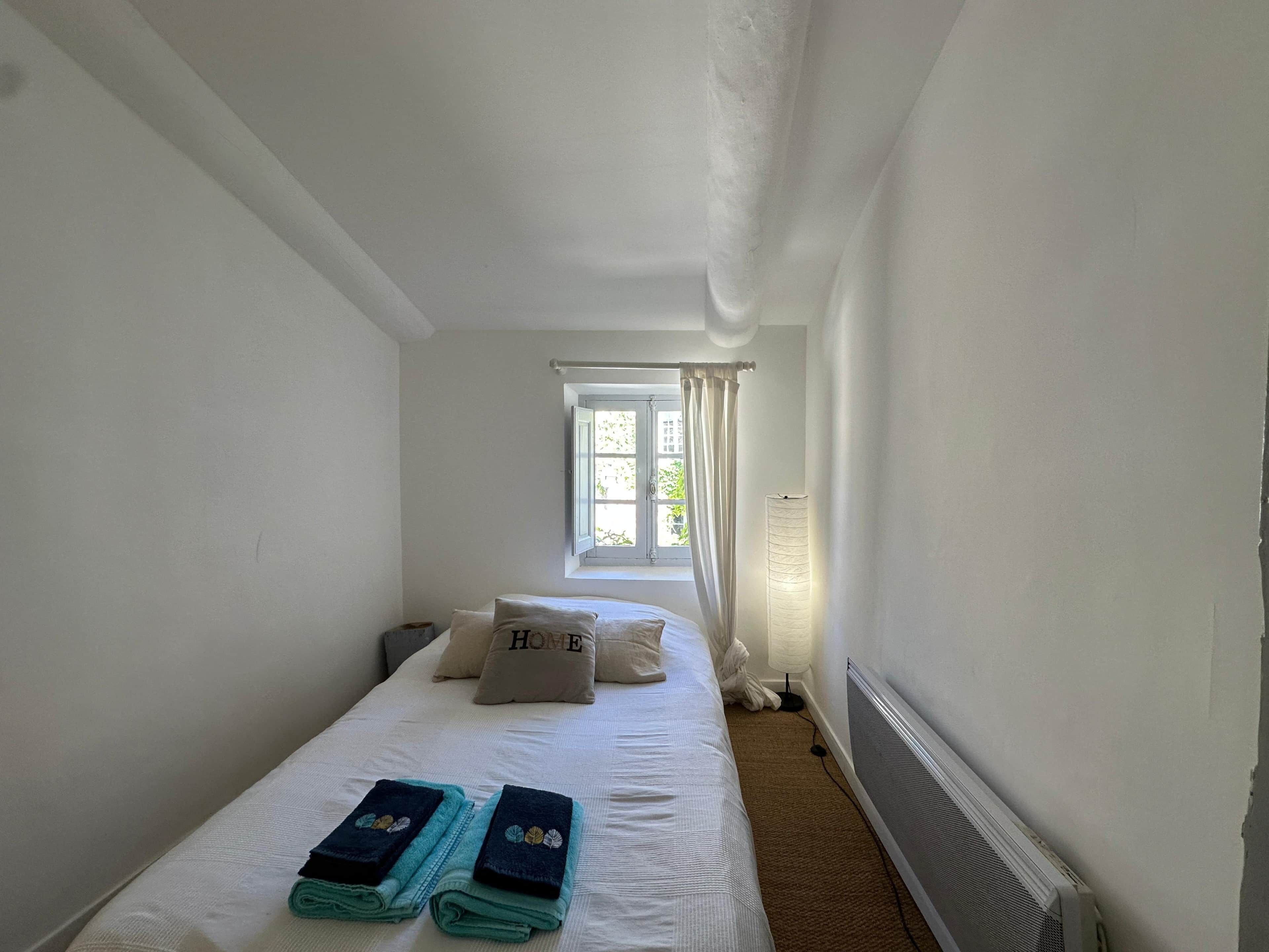 Ein schmaler, weiß getünchter Raum mit einem Einzelbett, einem Fenster mit Vorhängen und einer Heizung.
