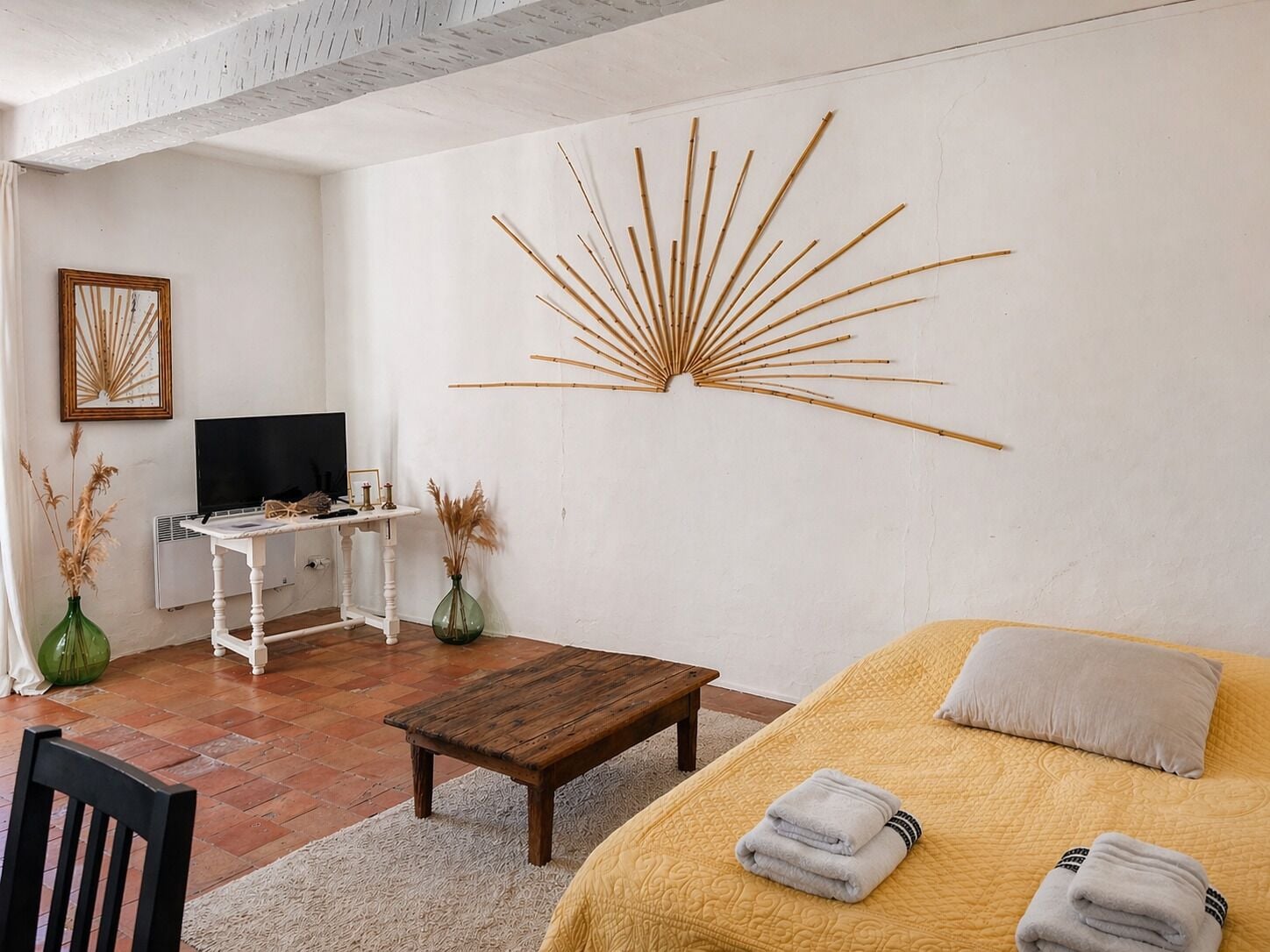 A room with a bed, a wooden coffee table, a TV on a white stand, a decorative wall piece, and two vases with dried plants.