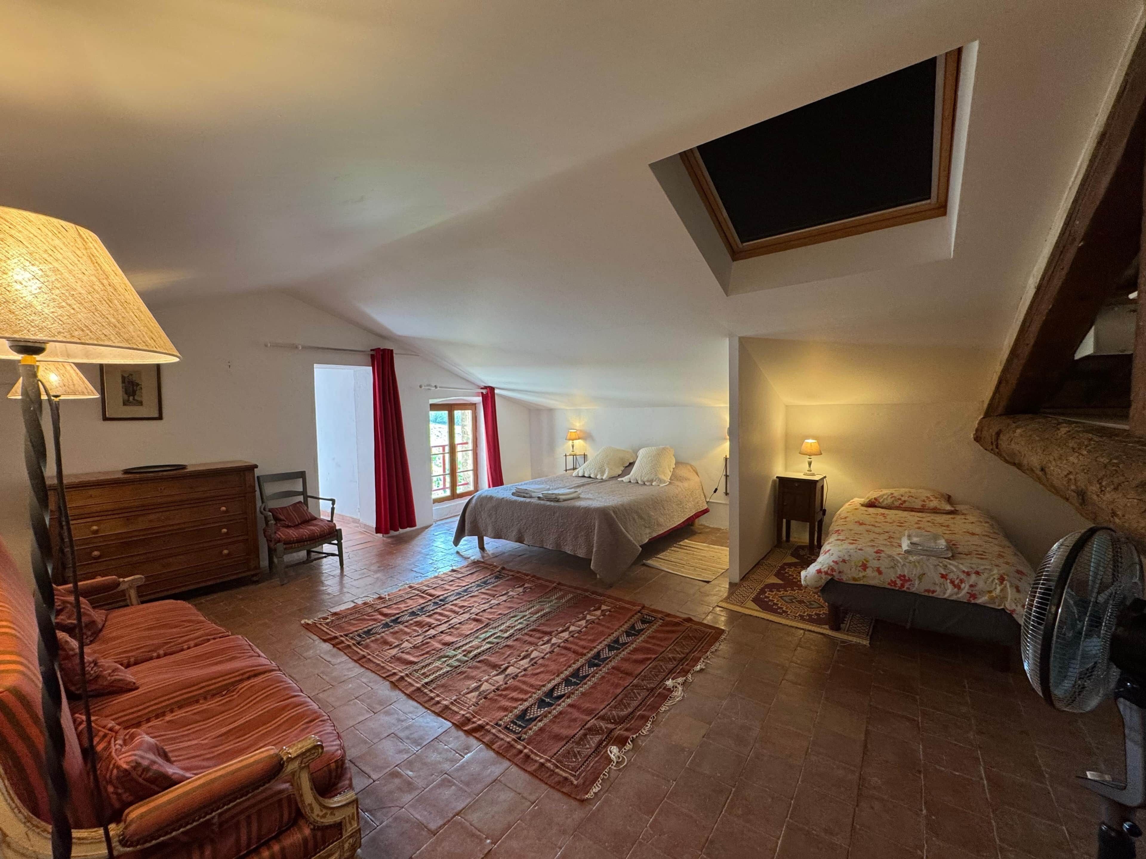 A spacious bedroom with a large bed, a sitting area with a sofa, a wooden dresser, and a fan.