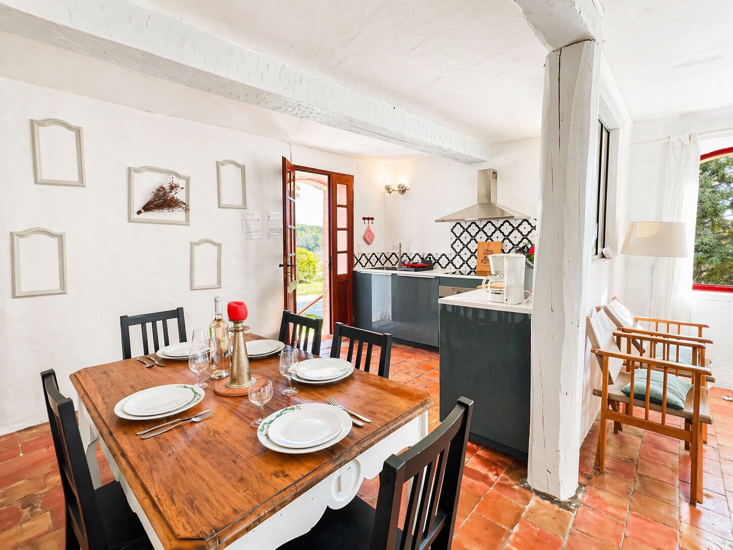 A dining area with a wooden table set for four, a kitchen with a tiled backsplash, and a view of greenery outside.