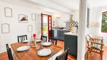 A dining area with a wooden table set for four, a kitchen with a tiled backsplash, and a view of greenery outside.