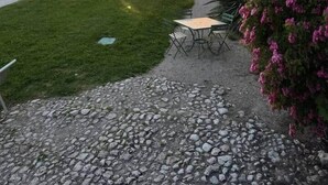 Sorapolkua, joka johtaa nurmialueelle, jossa on pöytä ja tuoleja. Taustalla näkyy kivirakennus, jonka seinää koristavat vaaleanpunaiset kukat, sekä auringonlasku.
