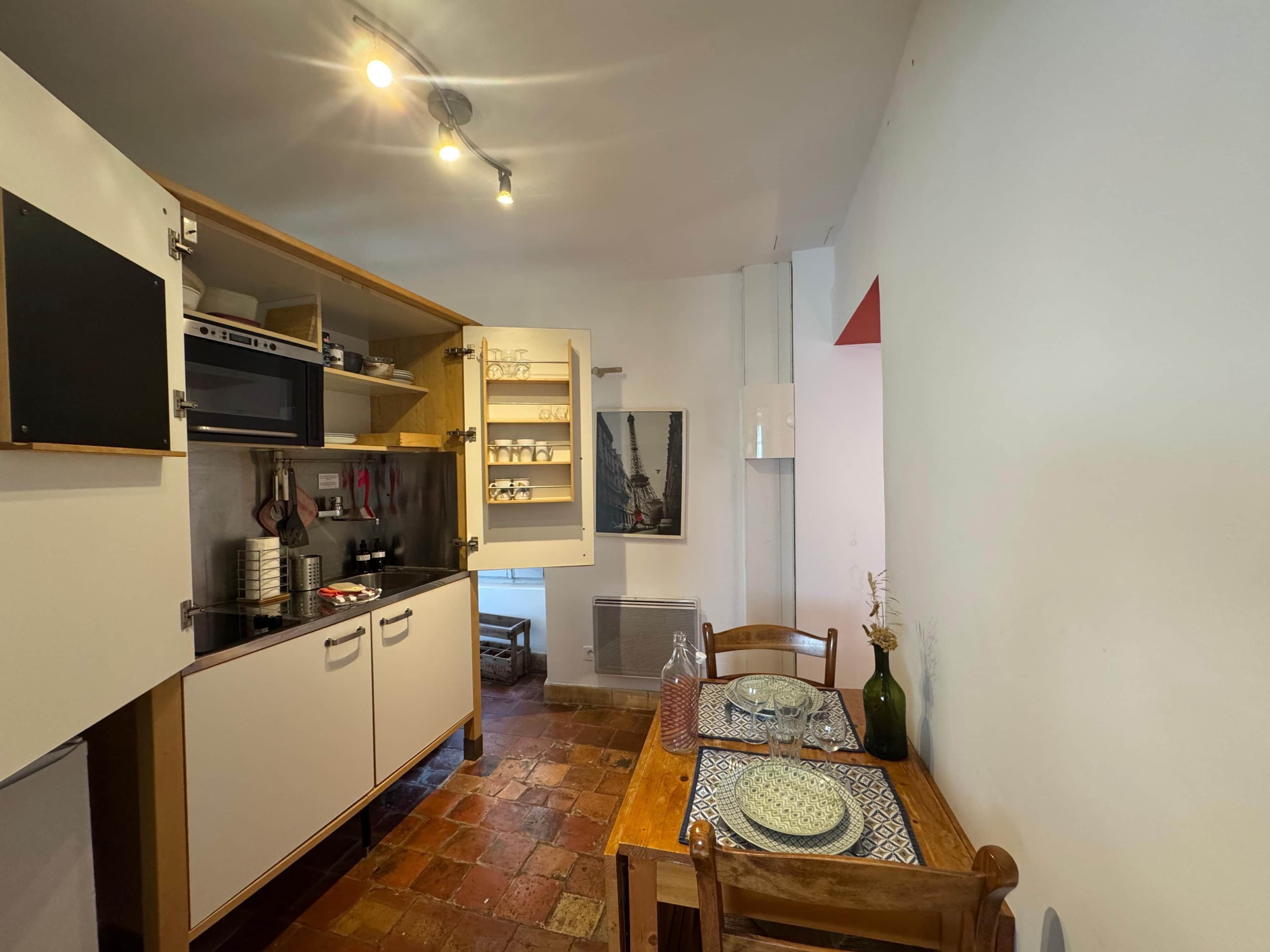 Une cuisine compacte équipée d’un four à micro-ondes, d’une cuisinière et d’un meuble ouvert où l’on peut voir la vaisselle et les ustensiles. Une table à manger, agrémentée de sets de table et d’un vase, est dressée pour deux personnes.