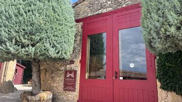 Un edificio de piedra con puerta roja, un cartel que indica bar y restaurante, y un gran árbol de hoja perenne al frente.