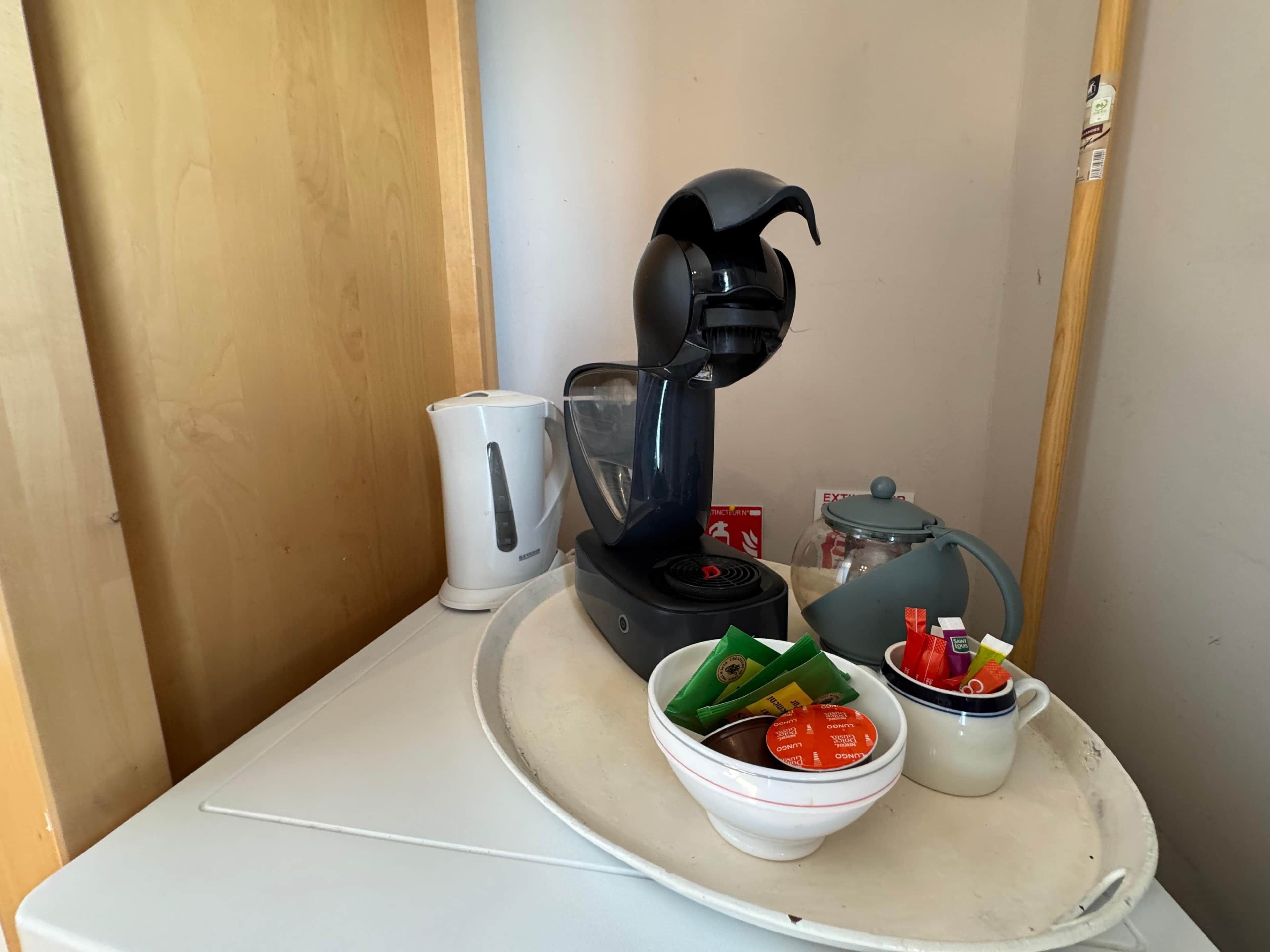 Un área de kitchenette con cafetera, hervidor, un tazón con bolsitas de té y una taza sobre una bandeja.