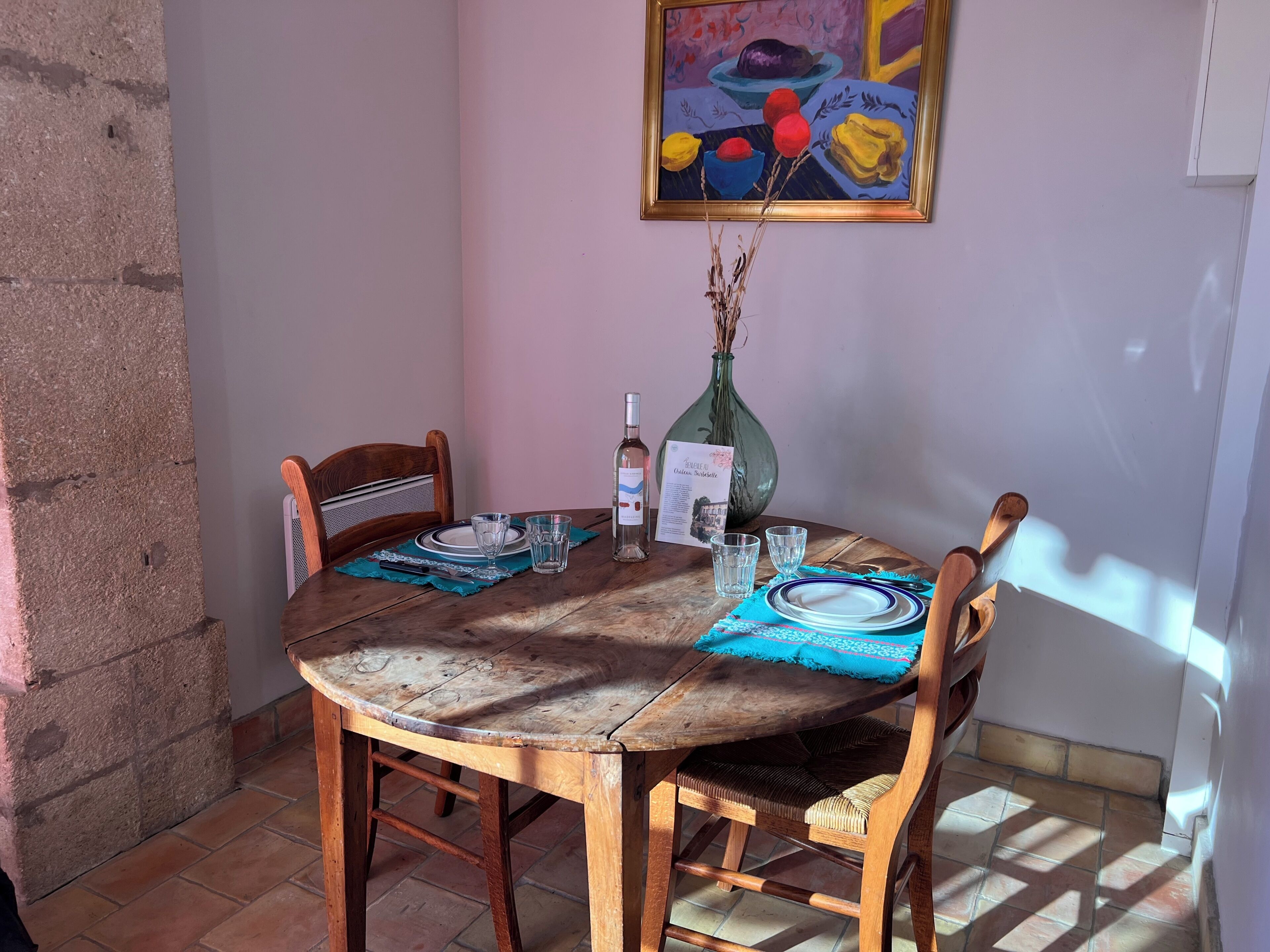 Una mesa de madera redonda con sillas, un jarrón con plantas secas, una botella y un menú sobre un mantel individual.