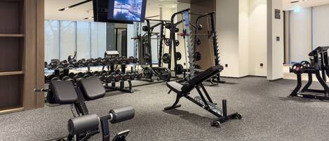 A gym with various exercise equipment, a television mounted on the wall, and a mirrored wall reflecting the equipment.