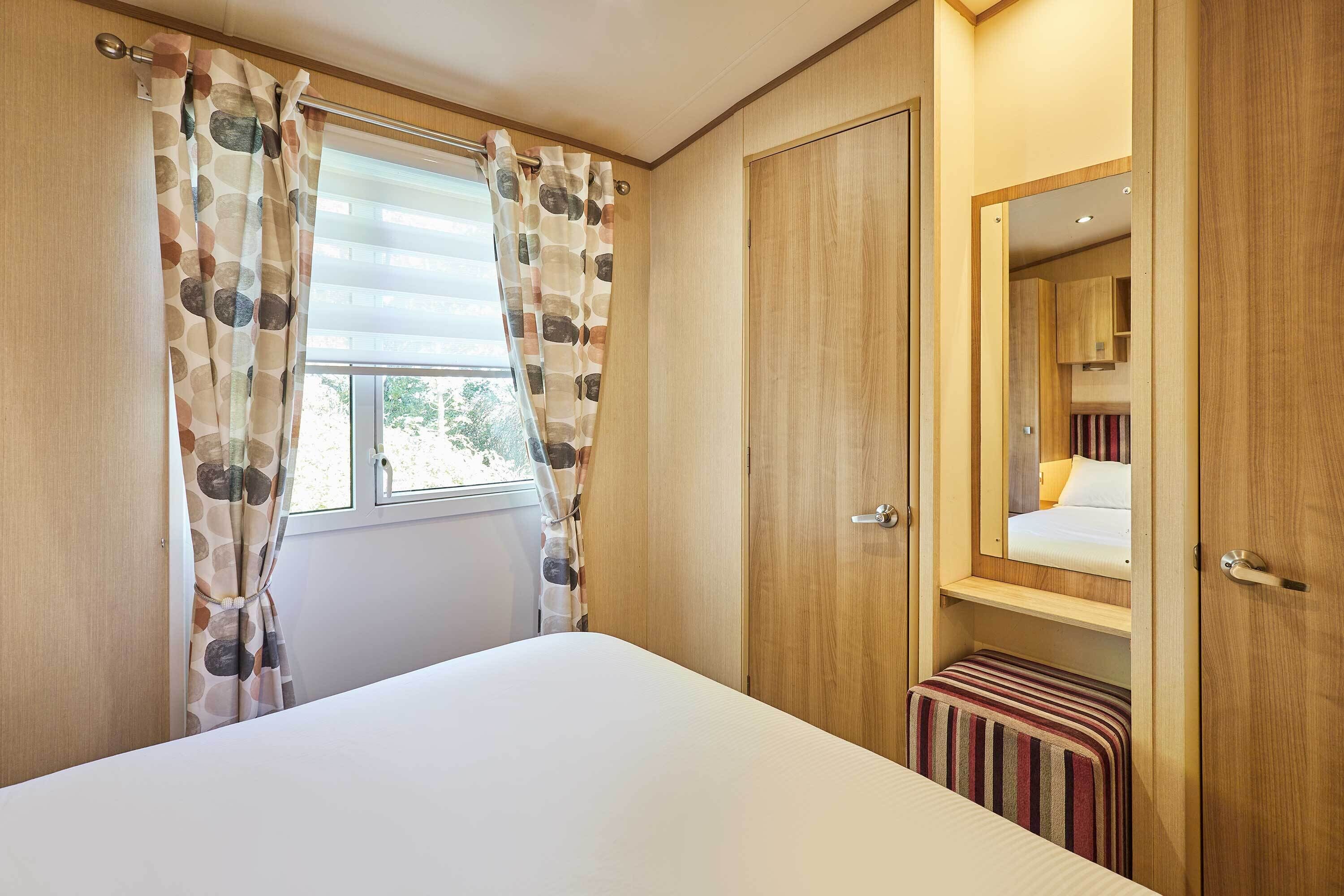 A bedroom with a bed, a window with curtains, a wooden wardrobe, and a small bench.
