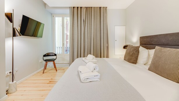A modern hotel room with a large bed, a flat-screen TV mounted on the wall, a chair, and a view of the outside through a window with curtains.