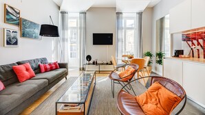 A modern living room with a sofa, coffee table, and chairs, a flat-screen TV, and a kitchen area with cabinets and a countertop.