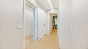 A hallway with white walls and wooden flooring, leading to a room with a chair and a table.
