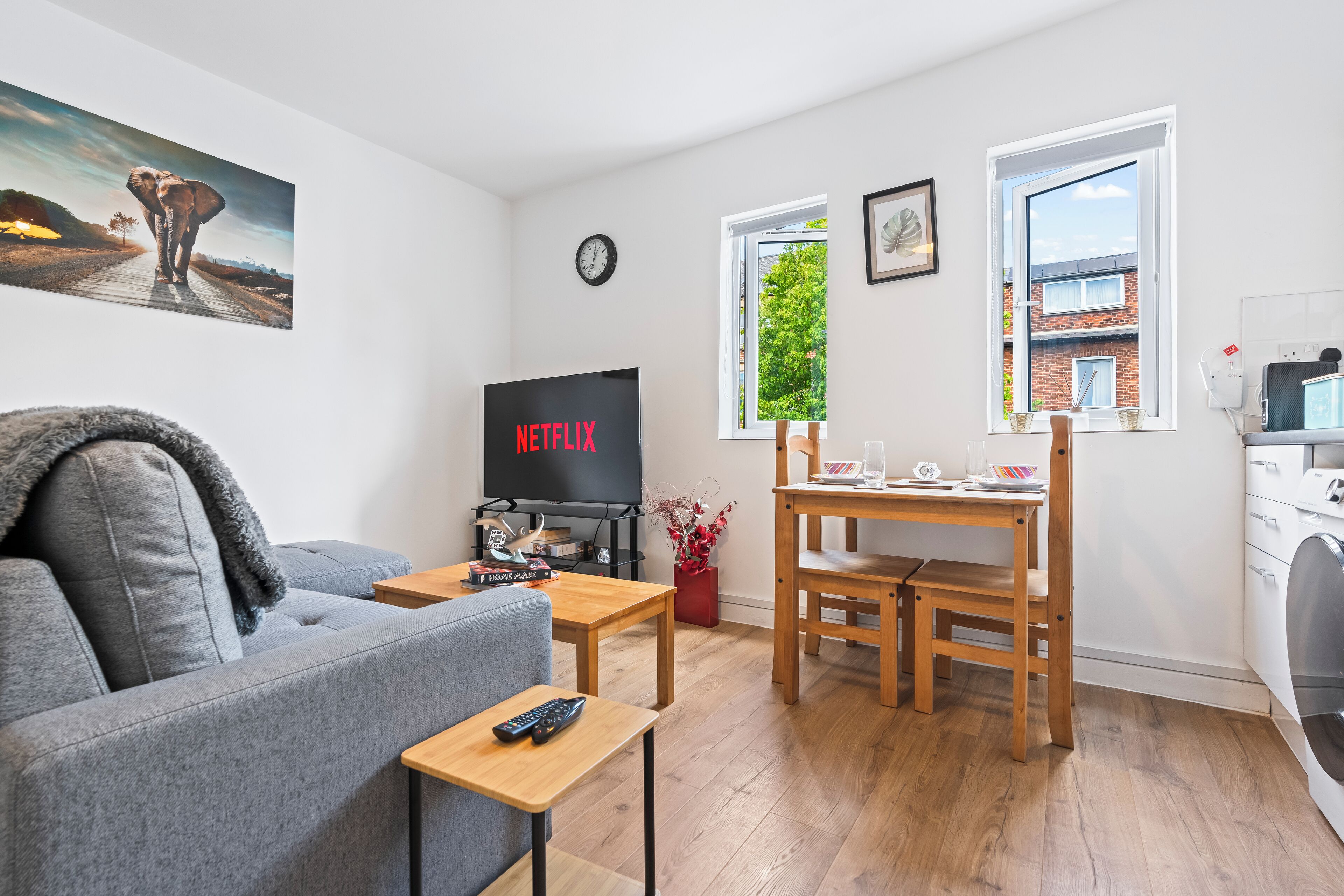 A modern living room with a grey sofa, a wooden dining table, a TV displaying 'NETFLIX', and a window with a view of greenery.