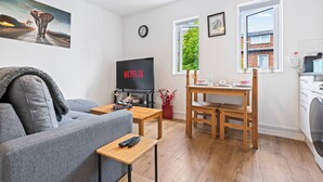 Un soggiorno moderno con un divano grigio, un tavolo da pranzo in legno, una TV che mostra 'NETFLIX' e una finestra con vista su del verde.