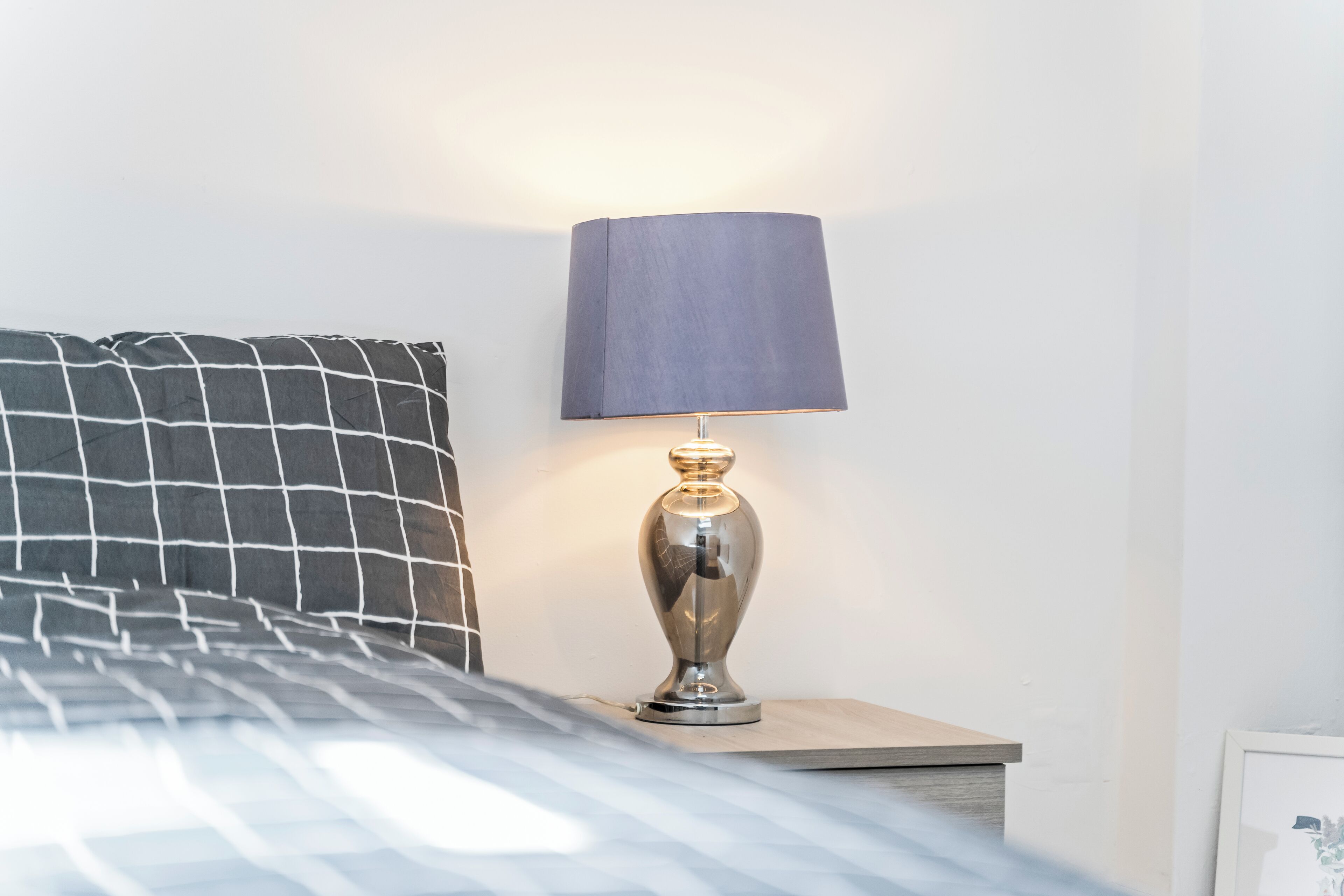 Ein ordentlich bezogenes Bett mit einem gemusterten Kopfkissen, eine moderne Lampe auf einem Nachttisch und ein gerahmtes Bild an der Wand.