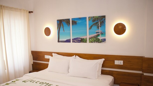 A hotel room with a bed, a headboard, a small table, and a wall-mounted picture of a beach scene. - Oshi Boutique  (Felidhoo)