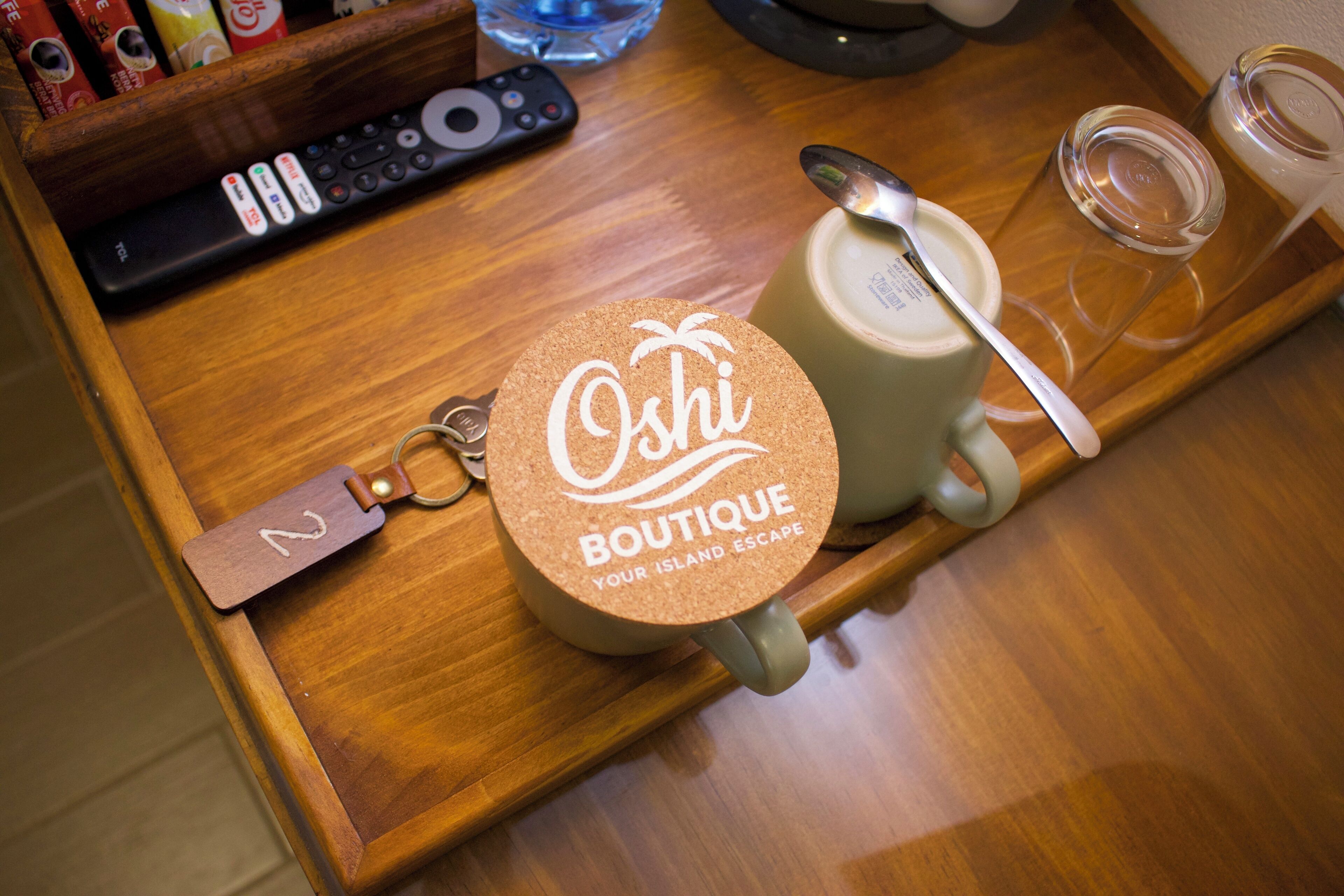A wooden tray with a cork coaster labeled "Oshi Boutique," a remote control, a spoon, a mug, and a glass jar.