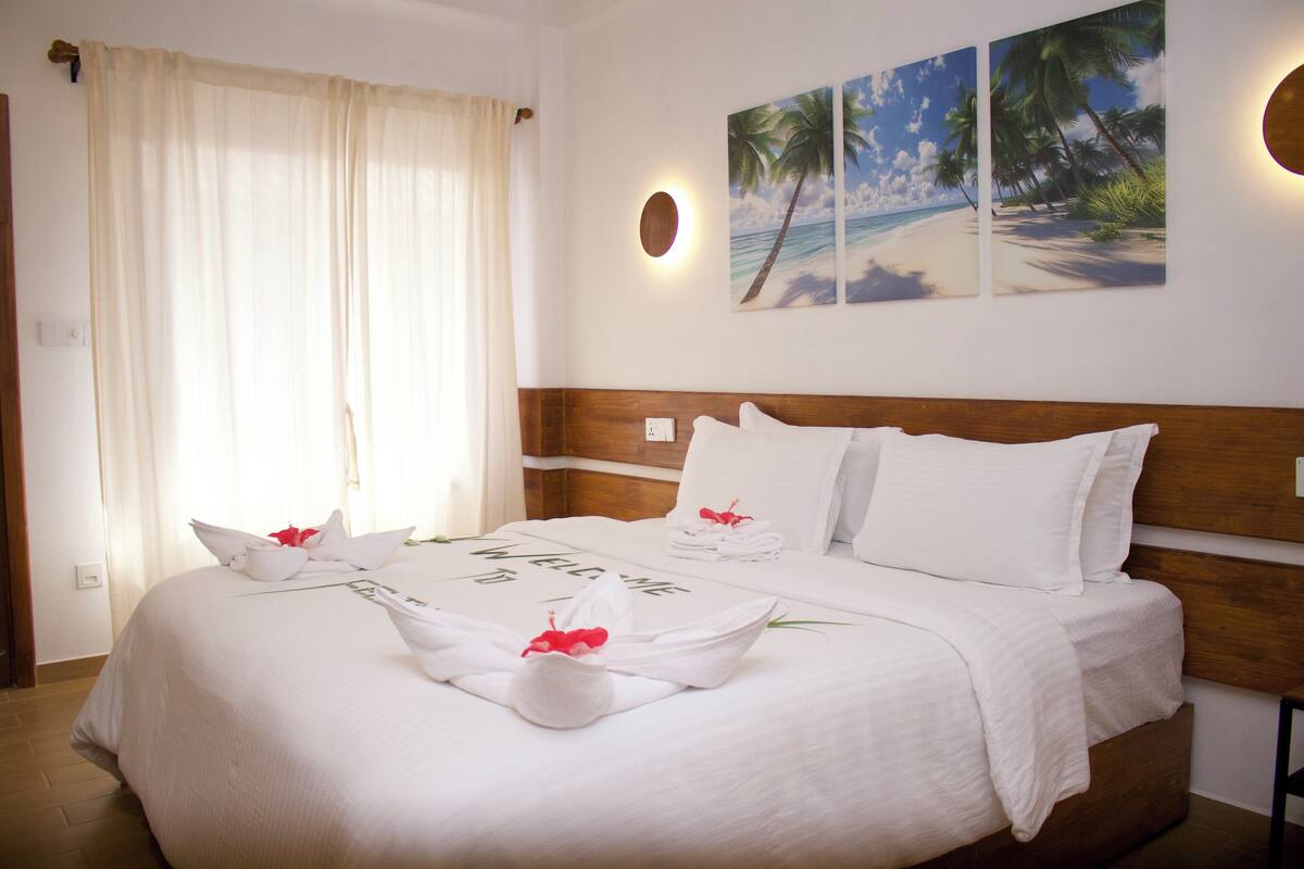 A neatly made bed with white linens and a welcome mat, a wooden headboard, and a wall with three tropical beach paintings.