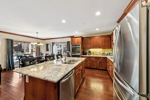A modern kitchen with a large island, stainless steel appliances, wooden cabinets, and a dining area with a view.