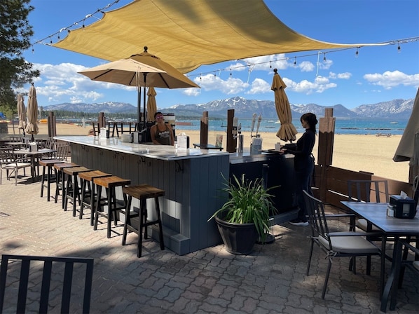 A beachside bar with outdoor seating, a counter, and a view of the beach and mountains.