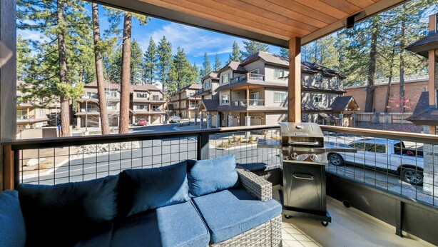 Un balcon avec un canapé en osier, un barbecue et une vue sur d’autres immeubles et des arbres.