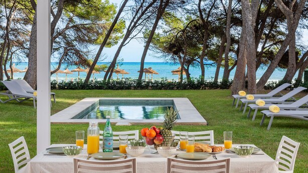 Une table à manger dressée avec des assiettes, des verres et un saladier de fruits, donnant sur une piscine et la mer.
