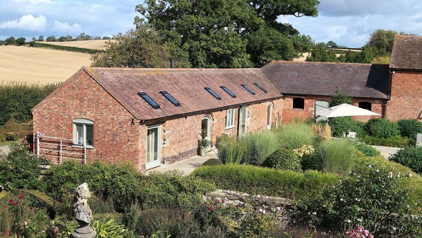 A brick building with multiple skylights, a garden with various plants and flowers, and a statue in the foreground. - Relaxing Retreat in Shropshire (Eaton Constantine)